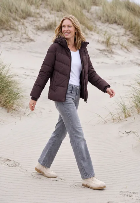Eine Frau in Steppjacke, Jeans und Sneakers spaziert lächelnd durch Dünen am Strand.