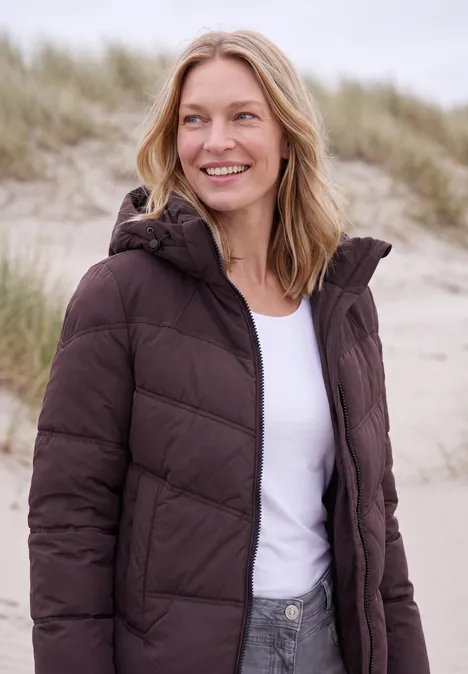 Eine lächelnde Frau in brauner Steppjacke und weißem Shirt am Strand vor Sanddünen.