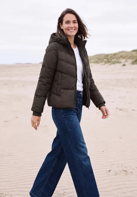 Frau in Winterjacke und Jeans spaziert am Strand, lächelt in die Kamera