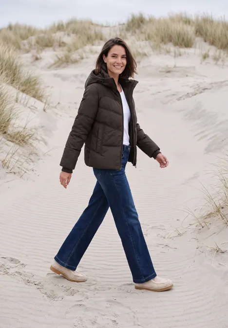 Eine lächelnde Frau in Daunenjacke und Jeans spaziert entspannt durch Dünen am Strand.