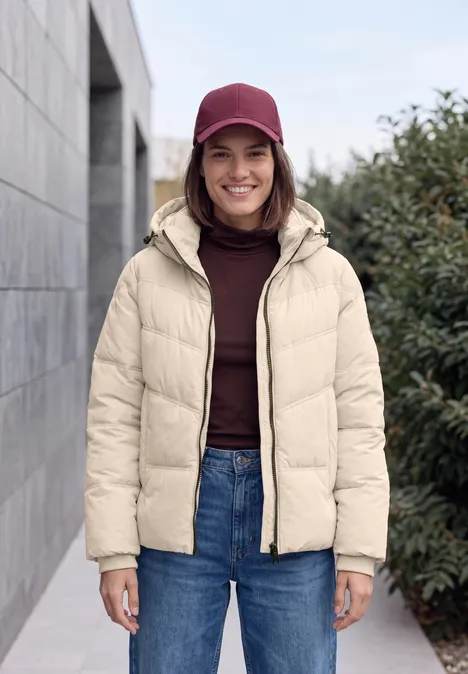 Junge Frau in beiger Daunenjacke und roter Mütze lächelt vor Hauswand und Pflanzen.