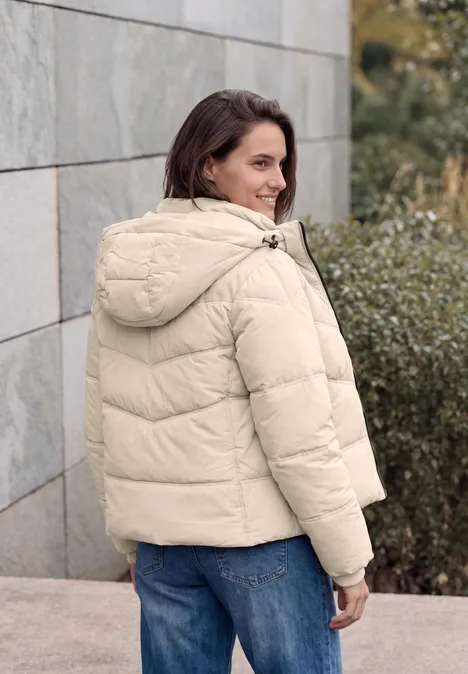 Frau in cremefarbener Steppjacke, lehnt an einer Steinwand und blickt lächelnd zurück.