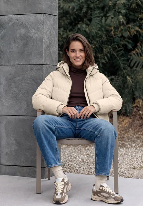 Junge Frau sitzt draußen auf Stuhl, trägt beige Daunenjacke, blaue Jeans und Sneaker, lächelt entspannt.