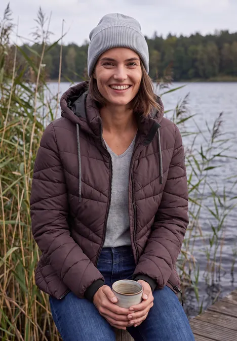 Veste matelassée à capuche et fermeture zip berry red Femme en doudoune et bonnet souriant assise sur un ponton au bord du lac, une tasse à la main.