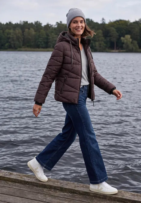 Veste matelassée à capuche et fermeture zip berry red Une femme souriante en jean, doudoune et bonnet marche sur une passerelle au bord d’un lac.