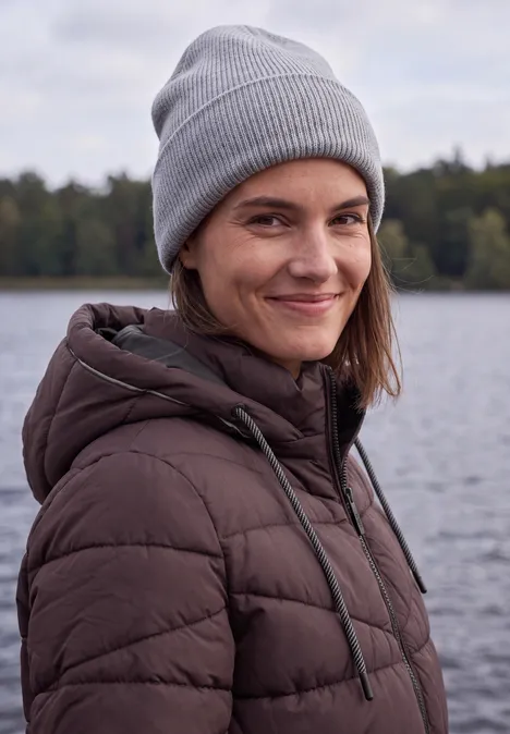 Veste matelassée à capuche et fermeture zip berry red Femme en bonnet gris et doudoune marron souriante devant un lac.