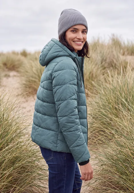 Frau in grüner Steppjacke und grauer Mütze steht in Dünenlandschaft und lächelt über die Schulter.