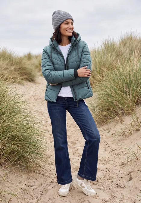 Frau in Winterjacke, Jeans, Mütze und Sneakers steht lächelnd zwischen Dünen am Strand