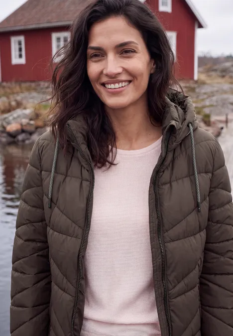 Lachende vrouw in een bruin winterjack en roze trui voor een rood huis aan het water.