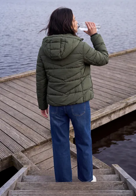 Person mit grüner Steppjacke und blauer Jeans trinkt am Holzsteg, Rückenansicht.