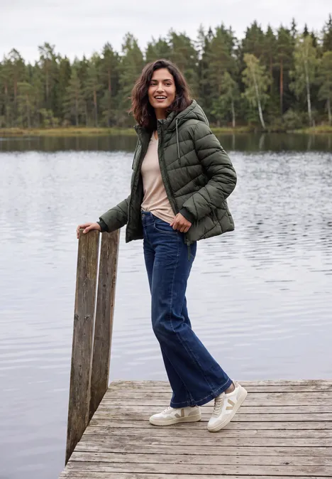 Frau mit grüner Steppjacke, Jeans und weißen Sneakern steht lächelnd auf einem Steg am ruhigen See.