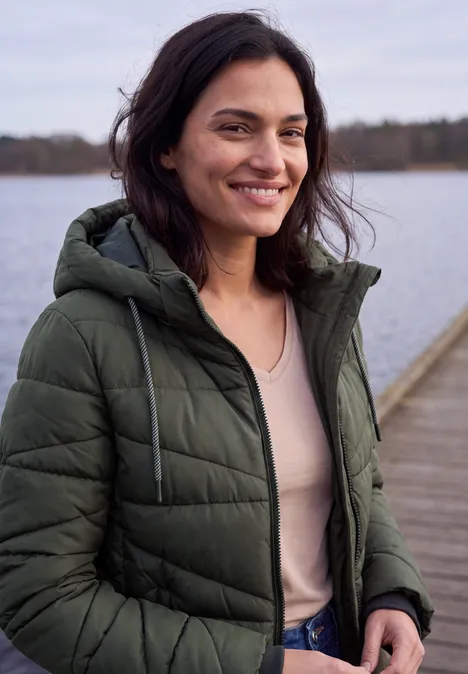 Lächelnde Frau in grüner Steppjacke steht auf einem Steg vor einem See.