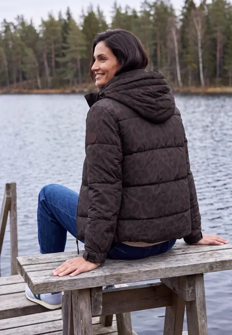 Frau sitzt lächelnd in Steppjacke auf Holzbank am See, umgeben von Wald und ruhigem Wasser.