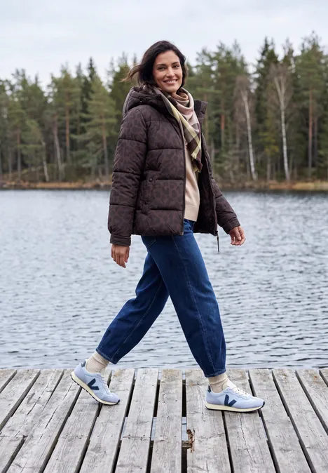 Frau in Winterjacke und Sneakern spaziert lächelnd auf einem Steg am See vor Waldkulisse.