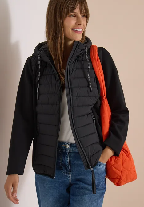 Femme en veste matelassée noire, jean et débardeur blanc, sac orange, sourire, fond neutre.