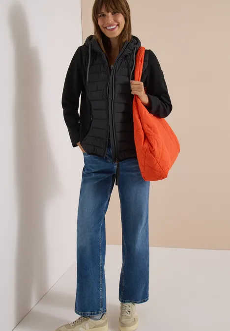 Femme en veste matelassée noire, jean large, baskets blanches et sac orange.