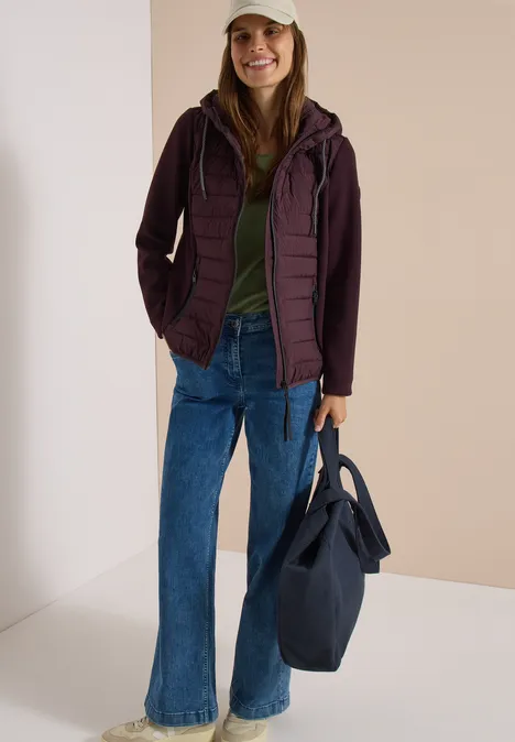 Une femme posant avec une veste matelassée, un t-shirt vert, un jean évasé, une casquette et un sac.