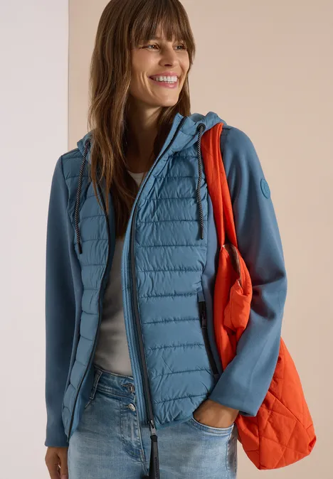 Femme souriante en veste matelassée bleu clair à manches Softshell, jean et sac matelassé orange.