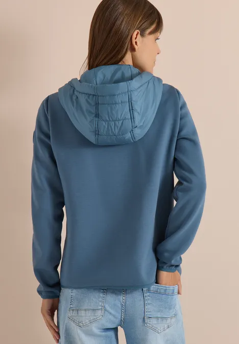 Femme vue de dos portant un sweat à capuche bleu matelassé et un jean sur un fond neutre.