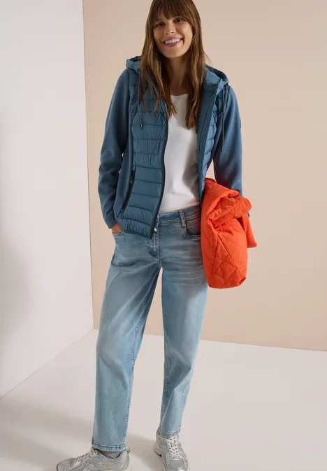 Femme décontractée en jean, t-shirt blanc, veste matelassée verte et sac orange, souriante.