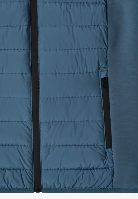 Gros plan sur une veste hybride bleue, face matelassée et poche zippée.