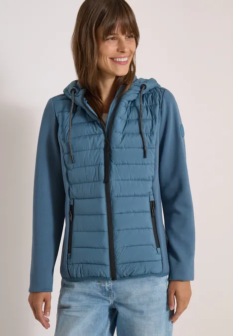 Femme portant veste à capuche bleue, devant matelassé et fermeture éclair, souriante.