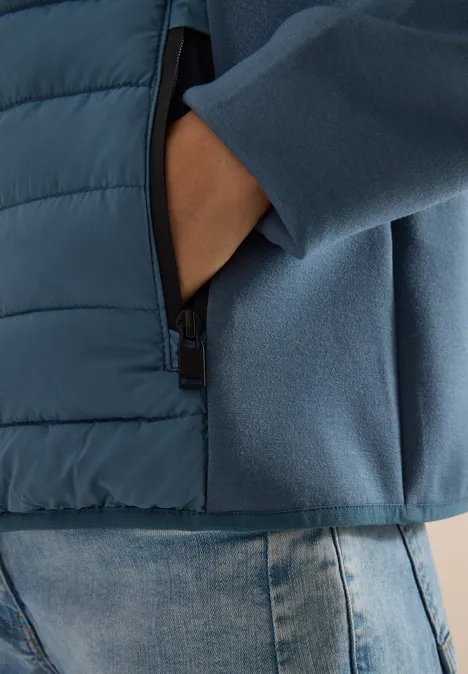 Main dans la poche zippée d'une veste bicolore bleue portée sur un jean.