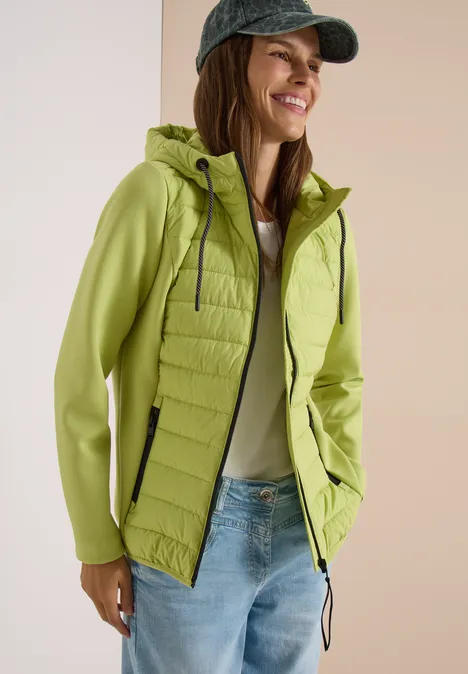 Femme souriante en veste à capuche vert clair et jean, vue de côté.