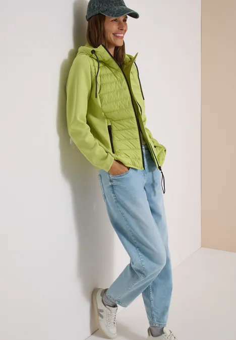 Une femme souriante appuyée contre un mur, vêtue d’une veste verte, d’un jean et d’une casquette.