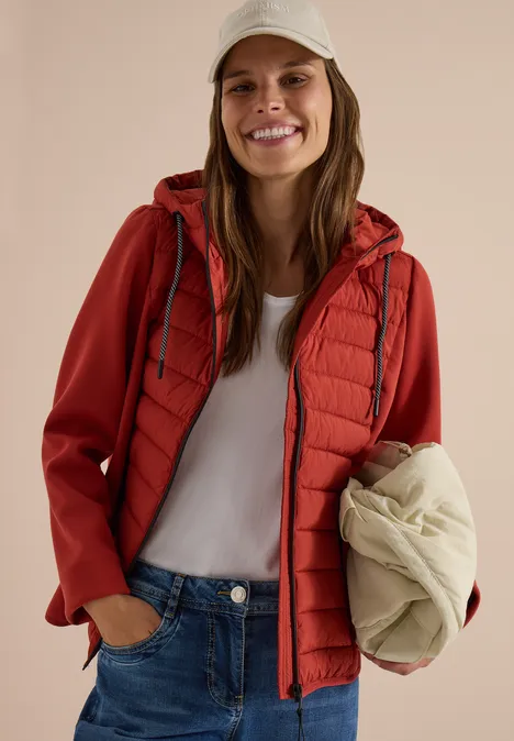 Femme souriante en veste rouge et t-shirt blanc, tenant une doudoune crème pliée.