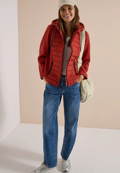 Femme portant une veste à capuche rouge, un gilet matelassé, jean bleu, baskets blanches et casquette.