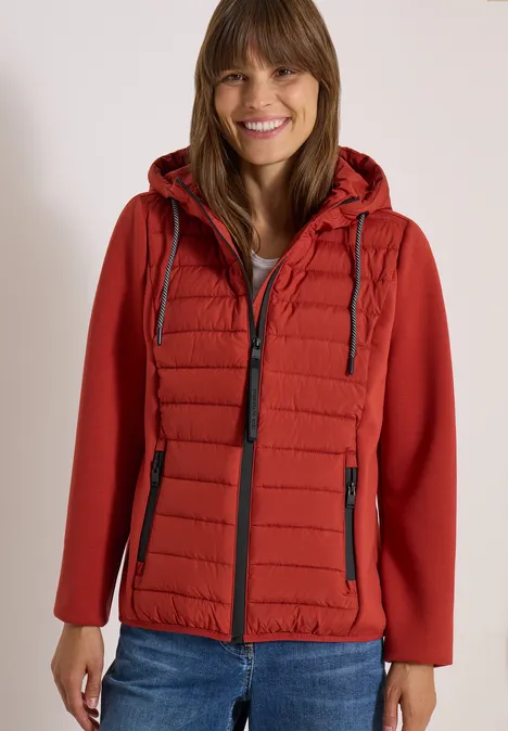 Femme souriante en veste matelassée rouge à capuche et jean devant un fond clair.