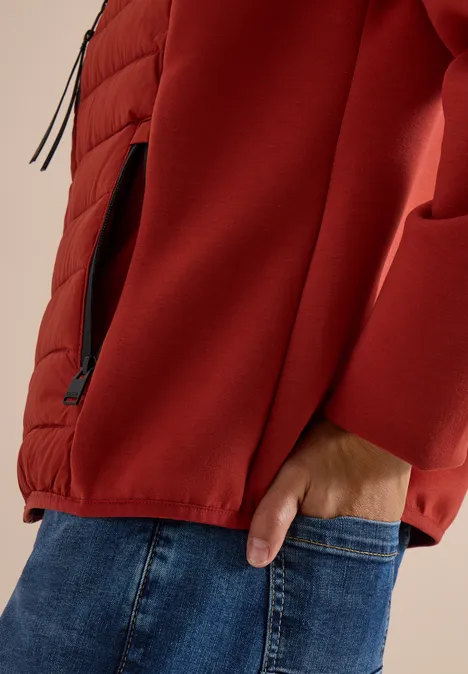 Haut du corps vêtu d’une veste rouge, main dans la poche de jean, poche zippée latérale visible.