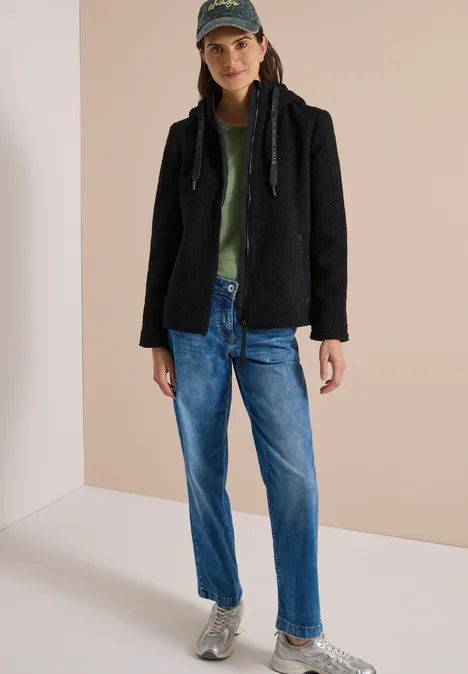 Junge Frau trägt schwarzen Kapuzenpullover, grünes Shirt, blaue Jeans und Sneaker, dazu Baseballcap.