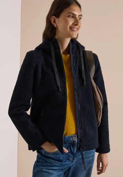 Femme souriante portant un blouson teddy bleu marine, un pull jaune et un jean, sac à l’épaule.