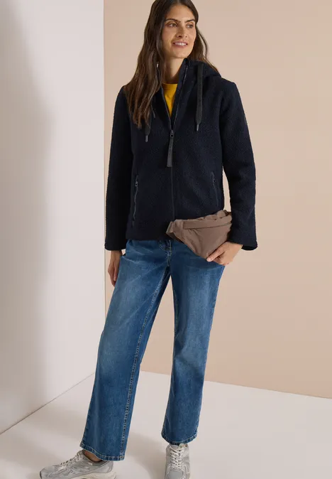 Femme en manteau bleu marine, jean large et pull jaune, tenant une pochette marron devant un fond neutre.