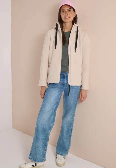 Frau in beiger Fleecejacke, grünem Shirt, weiten Jeans, weißen Sneakern und pinker Cap vor hellem Hintergrund.