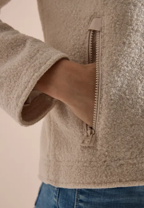 Eine Hand steckt in der Reißverschlusstasche einer beigen, flauschigen Jacke.