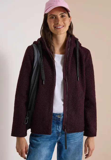 Femme en veste polaire bordeaux, T-shirt blanc, jean et casquette rose, avec sac à dos, souriante.