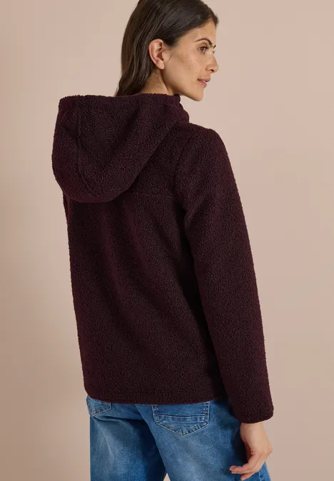 Femme de profil portant une veste teddy bordeaux à capuche et un jean.