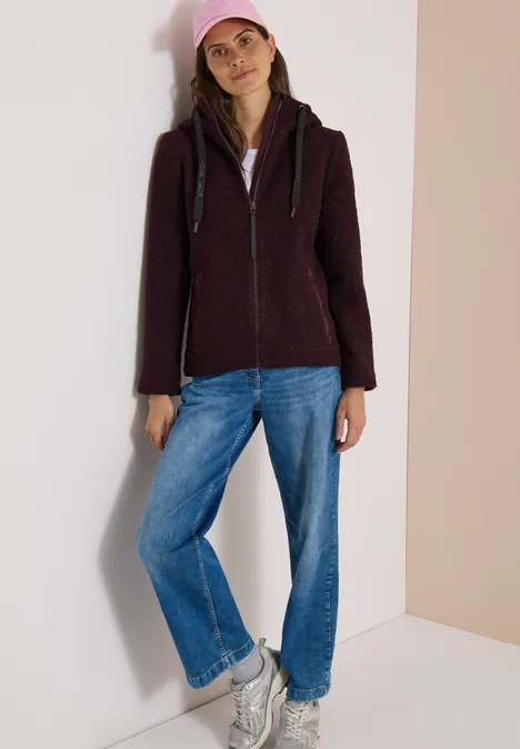 Femme en sweat à capuche sombre, jean bleu et baskets blanches appuyée contre un mur clair.