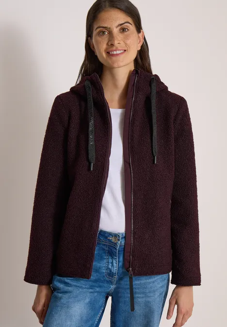 Jeune femme souriante portant une veste à capuche violette foncée et un jean.