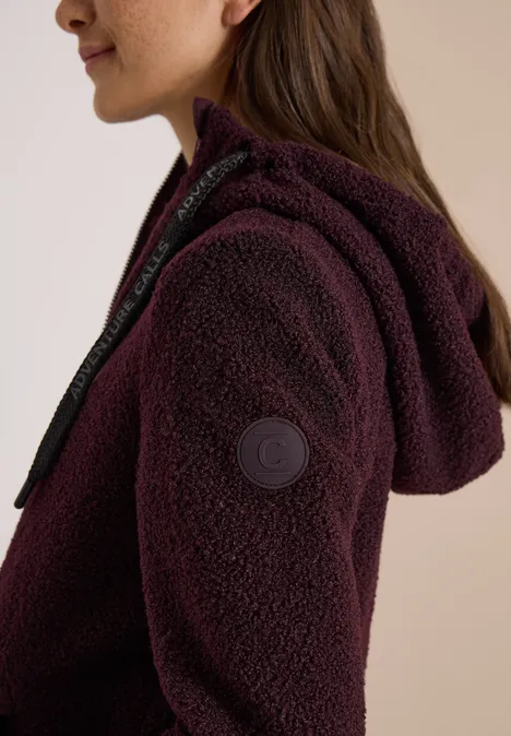 Femme portant une veste teddy polaire violette à capuche, zip et logo de marque sur la manche.