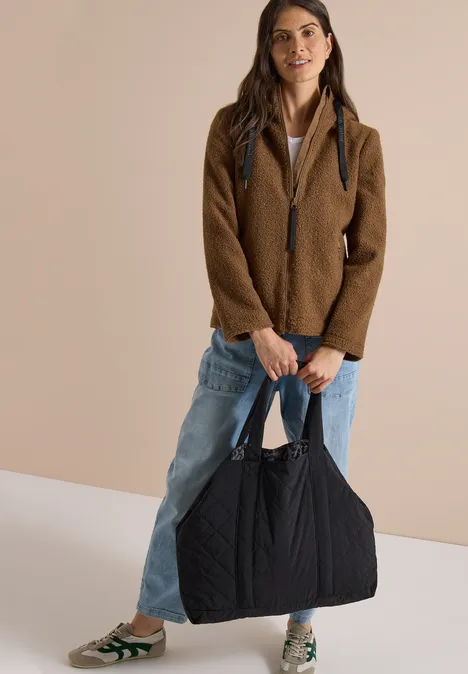 Frau in brauner Jacke und Jeans hält große, schwarze Stepp-Tasche und lächelt.