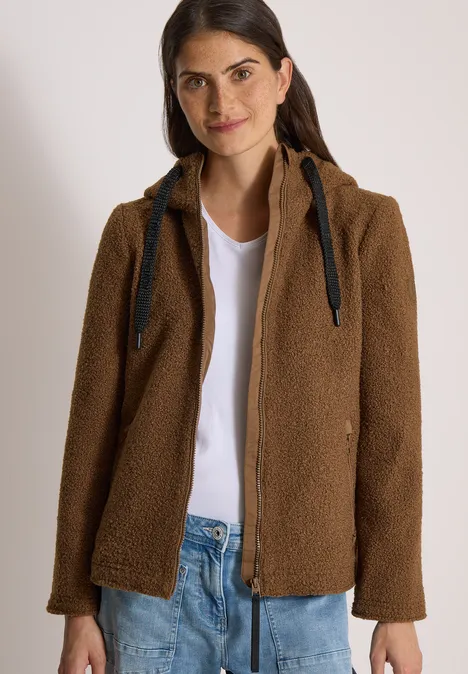 Frau mit brauner Teddy-Jacke, weißem Shirt und Jeans vor hellem Hintergrund.