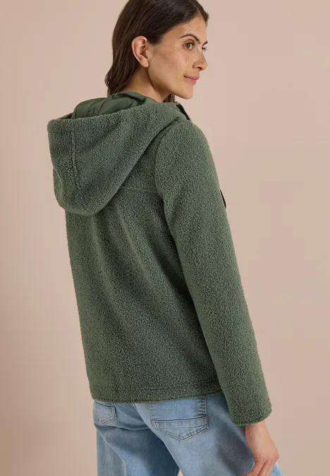 Femme vue de dos portant une veste teddy verte à capuche et un jean, fond beige.