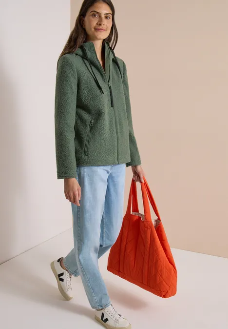 Femme portant une veste à capuche verte, jean clair, baskets blanches et sac en tissu orange sur fond neutre.
