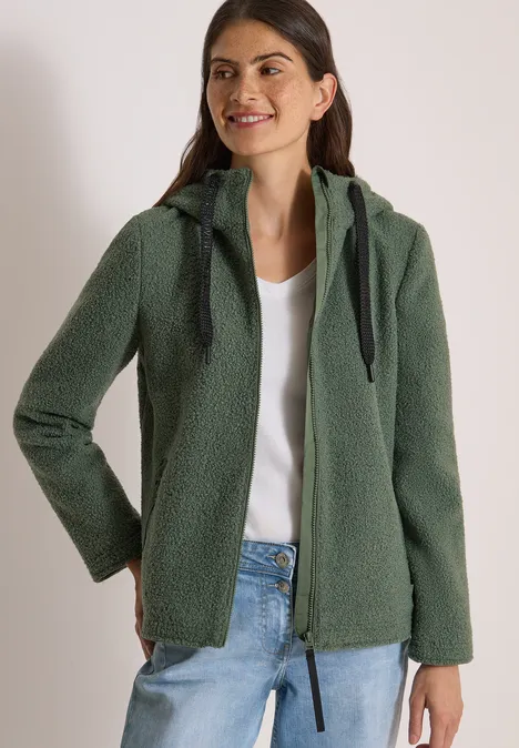 Femme souriante en veste en polaire vert à capuche, chemisier blanc, mains dans les poches.
