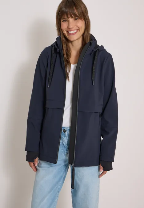 Frau mit Jeans und weißem Shirt lächelt und trägt eine marineblaue Kapuzenjacke.