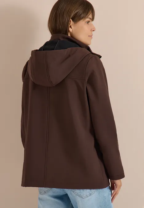 Femme de dos en veste à capuche marron et jean devant un fond beige.